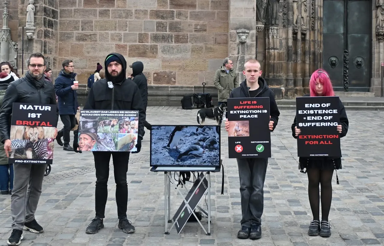 Protest in Germany: five activists in front of a church hold signs such as "Natur ist brutal", "Life is suffering, extinction is peace" and "End of existence = end of suffering", next to a screen showing a video